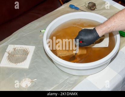 Lavare le parti in plastica di una ventola di scarico della cucina molto sporca in una soluzione acquosa di idrossido di sodio. Un uomo lavora in guanti protettivi e torace Foto Stock