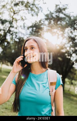 Ragazza studentesca adolescente che indossa una t-shirt acquamarina, facendo una telefonata alla sua famiglia nel parco. Retroilluminazione del sole Foto Stock