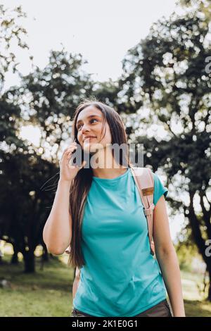 Ragazza studentesca adolescente che indossa una t-shirt acquamarina, facendo una telefonata alla sua famiglia nel parco. Retroilluminazione del sole Foto Stock