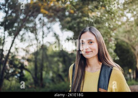 Sorridente studentessa universitaria che indossa un maglione giallo e uno zaino nella strada del parco del campus. Sorriso innocente Foto Stock