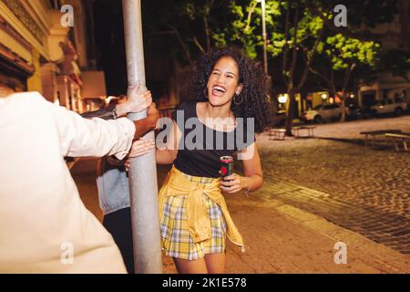 Amici che giocano intorno ad un palo di notte. Gruppo di giovani amici allegri che ridono e si divertono in città. Ritrovo multiculturale dei giovani Foto Stock