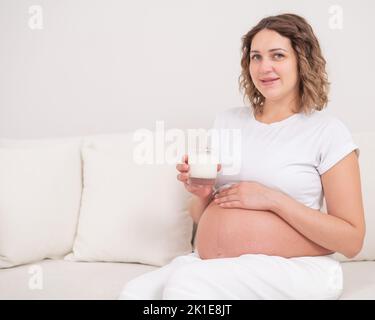 Una donna incinta siede su un divano bianco e beve latte. Foto Stock