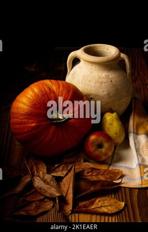 Un colpo verticale di una zucca, una caraffa di argilla e frutta su una superficie di legno con foglie secche Foto Stock