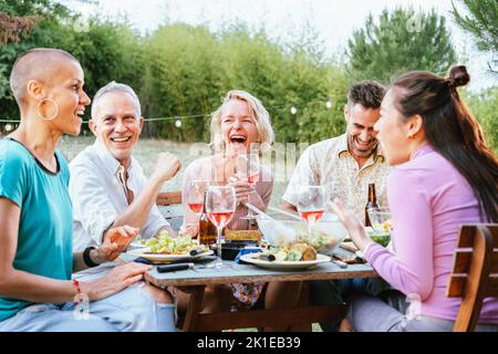 Gruppo di persone multietniche adulte che ride e si divertono a tenere bicchieri di vino durante la cena al cortile. Concetto di stile di vita. Foto Stock