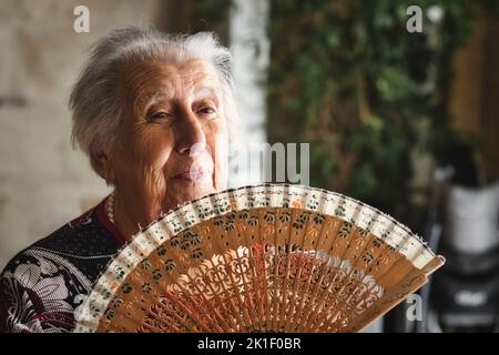 Una vecchia donna con i capelli bianchi all'interno che ondola un ventilatore tradizionale di legno per mantenersi fredda in estate Foto Stock