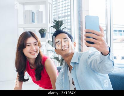 Felice bell'uomo asiatico e giovane bella donna selfie mentre si siede vicino alla finestra di vetro nel soggiorno bianco in alto edificio. Coppia allegra t Foto Stock