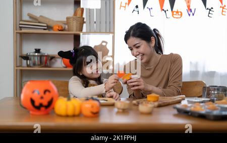 Felice famiglia sorridente madre e figlia facendo Halloween decorazioni in casa insieme, mentre si siede al tavolo di legno. Foto Stock