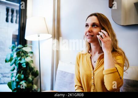 giovane donna bionda con camicia gialla seduta in una caffetteria e che parla per telefono Foto Stock