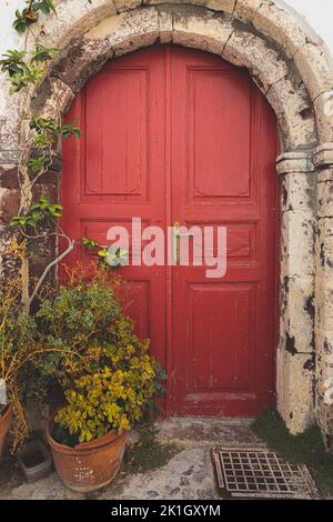 Particolare di una tradizionale e decorativa porta rossa sotto un arco di pietra nel villaggio della città vecchia di Oia, sull'isola greca di Santorini, Grecia. Foto Stock