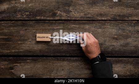 Mano di un uomo d'affari con un elegante orologio che dirige un origami fatto barca di carta tirando un piolo di legno con segno di Seguici scritto su di esso. Vista dall'alto Foto Stock