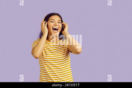 Donna positiva in cuffie ascolta la musica e canta fortemente isolato su sfondo viola pastello. Foto Stock