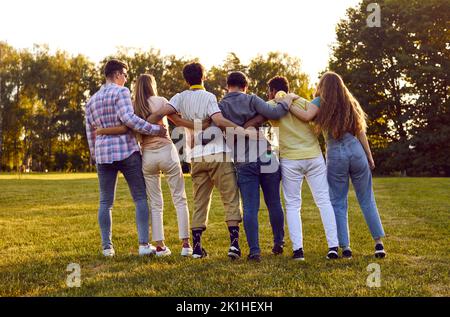 Vista posteriore di diversi amici in piedi nel verde parco estivo e abbracciarsi l'un l'altro Foto Stock