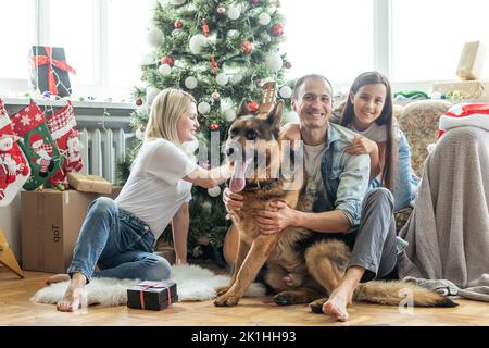 Ragazza eccitata e la sua famiglia seduta sul pavimento vicino albero di natale e sorridente. Famiglia durante Christmastime Foto Stock