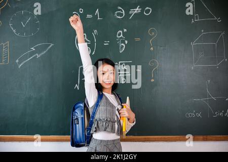 Istruzione Torna a scuola. Ragazza asiatica della scuola in uniforme sulle braccia alzate della classe per il successo finito compiti a casa, il bambino primario è in piedi davanti al cl Foto Stock