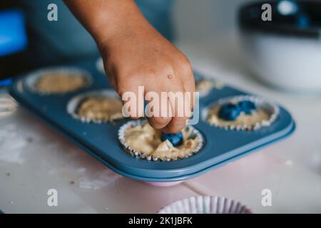 La mano di un ragazzo che mette mirtilli sulla pasta già divisa in forme di cottura per le torte di vacanza. Bambini, decorazione, carta da imballaggio, tessuto. Foto Stock