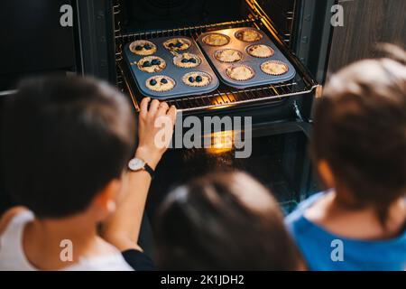 Madre con i suoi due figli mettere cupcake fatti in casa nel forno per cuocere. Dolcetti fatti a mano per le vacanze. Stile di vita delle persone. Famiglia felice, infanzia. Foto Stock