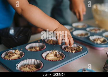 Le mani della ragazza cute che mettono le gocce di cioccolato sulla pastella grezza del muffin nelle teglie. Ricetta passo dopo passo Foto Stock