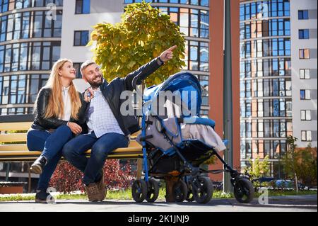 Famiglia felice seduta insieme con scivolando in bambino di pram blu. Donna sorridente che tiene le chiavi, uomo che indica un nuovo appartamento nel nuovo quartiere della città. Moderni edifici residenziali sullo sfondo. Foto Stock