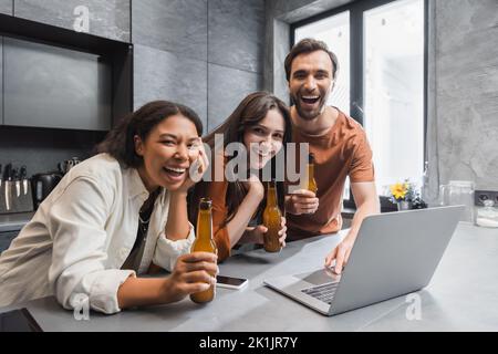amici multietnici felici tenendo bottiglie di birra vicino gadget e ridendo in cucina Foto Stock