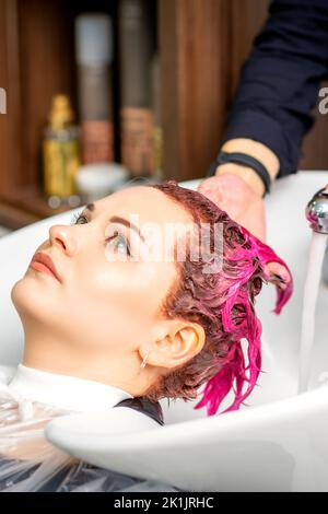 Lavaggio capelli tinti femmina. Una giovane donna caucasica che ha i capelli lavati in un salone di bellezza. Il parrucchiere professionale lava la vernice di colore rosa dai capelli di un cliente Foto Stock