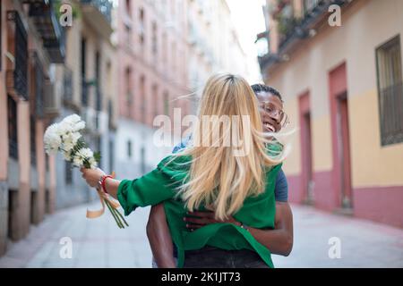 Concentratevi su amici multietnici che abbracciano in modo effusivo la strada, mentre si tiene un bouquet di fiori Foto Stock