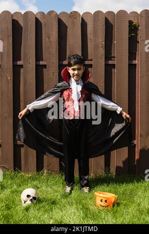 lunghezza intera del ragazzo asiatico in costume di vampiro che tiene il mantello vicino al cranio e secchio di halloween sull'erba Foto Stock