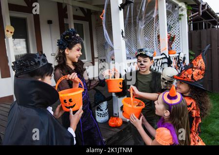 Bambini multietnici positivi che tengono secchi con caramelle mentre festeggia Halloween in cortile Foto Stock