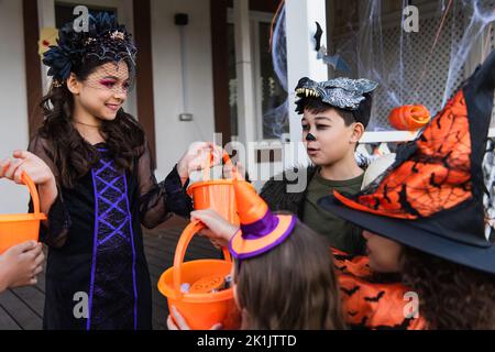 Capretto sorridente che tiene i secchi vicino agli amici interracial nei costumi di Halloween all'aperto Foto Stock