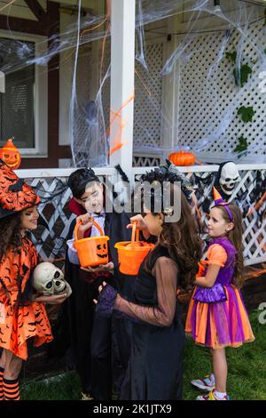 Bambini premiati sorridenti in costumi di Halloween guardando secchi con caramelle vicino amici interrazziali all'aperto Foto Stock