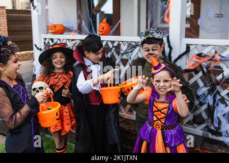 Bambini interrazziali in costumi di Halloween tenendo secchi e giocando all'aperto Foto Stock