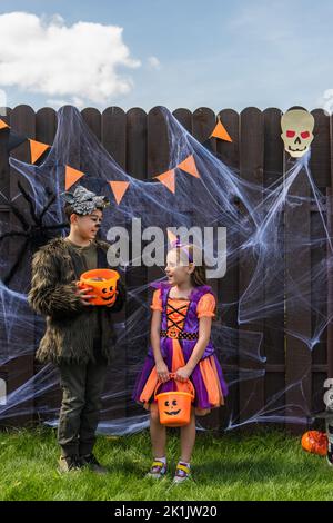 Sorridente bambini interracial in costumi di Halloween che tengono i secchi con le caramelle vicino al ragnatela e l'arredamento sulla recinzione all'aperto Foto Stock