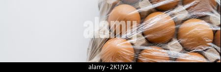 Vista dall'alto delle uova marroni sotto il cellofano in scatola di cartone su sfondo bianco, banner Foto Stock