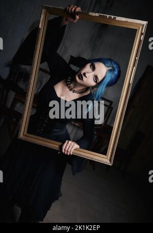 Ritratto in interni di bella goth ragazza in abito nero. Donna gotica dai capelli blu tiene in mano una cornice Foto Stock