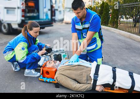 Paramedici EMS che eseguono massaggio cardiaco a torace chiuso e respirazione artificiale per l'uomo maturo ferito che si trova inconscio sulla barella medica nea Foto Stock