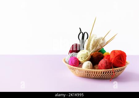 Rosso, giallo, bianco, viola, marrone, palle verdi di filo in un cesto beige con aghi da maglieria in bambù e forbici su fondo lilla Foto Stock