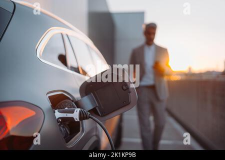 Primo piano della ricarica di auto elettriche, uomo d'affari in piedi in background e in attesa. Foto Stock