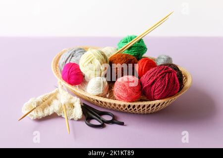 Rosso, giallo, bianco, viola, marrone, palle verdi di filo in un cesto beige con aghi da maglieria in bambù e forbici su fondo lilla, giacitura piatta Foto Stock