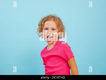 Ritratto di felice bambina con occhi blu con capelli ricci che guardano, mostrando la lingua, grimacing teasing su sfondo blu. Foto Stock