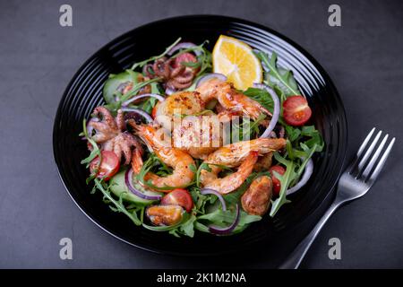 Insalata con frutti di mare, rucola, pomodori, cetrioli, cipolla rossa e limone su un piatto nero. Sfondo nero, primo piano. Foto Stock