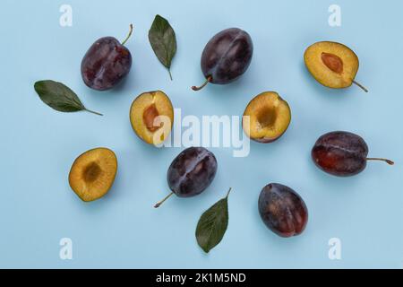 prugne fresche mature e foglie verdi su sfondo blu, giacitura piatta, vista dall'alto, modello di frutta intera e metà, cibo, sana alimentazione biologica, raccolta co Foto Stock