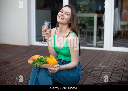 Ritratto di giovane felice bellissima donna seduta su un gradino di veranda in legno, tenendo un bicchiere di vino bianco, verdure tagliate. Foto Stock