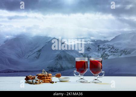 Design per biglietti d'auguri. Bicchiere con VIN brulé, torte, biscotti, miele e decorazioni natalizie sul tavolo, sullo sfondo delle montagne innevate d'inverno Foto Stock