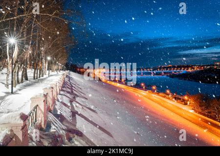 Vista della città invernale notturna coperta di neve e la strada con tracce di fari di auto dal parco cittadino. Nevicate. Foto Stock