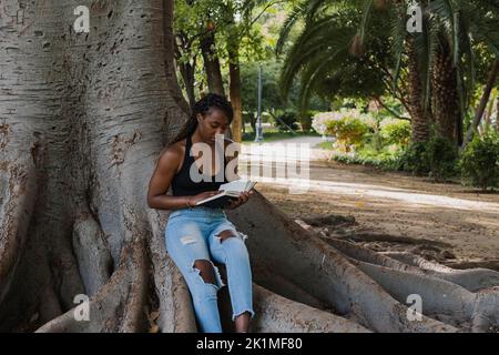 GIOVANE AFROAMERICANA SI SIEDE RILASSANDOSI ALL'OMBRA DI UN ALBERO CHE LEGGE UN LIBRO Foto Stock