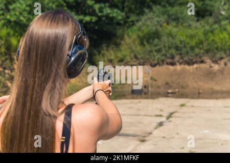 vista posteriore di una ragazza dai capelli lunghi che spara un bersaglio con una pistola sul campo di tiro in una giornata di sole. Foto di alta qualità Foto Stock