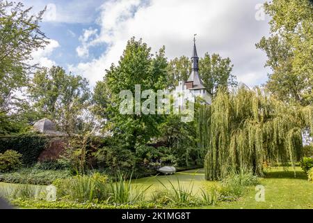 Paesaggio rurale olandese con ruscello circondato da erba verde e alberi lussureggianti, muro di mattoni al di fuori del castello Kasteel Groot Buggenum, torre campanaria sullo sfondo Foto Stock