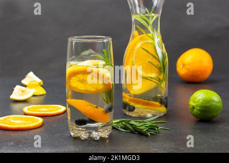 Fette di arancia tagliate e rametto di rosmarino sul tavolo. Bevanda estiva infusa con arance, mirtilli e rosmarino in bicchiere e bottiglia. Arancio e lime Foto Stock