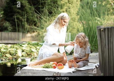 Ritratto di famiglia seduto su plaid bianco sul bordo dello stagno. Giovane donna che versa il latte in vetro per bambina figlia. Foto Stock