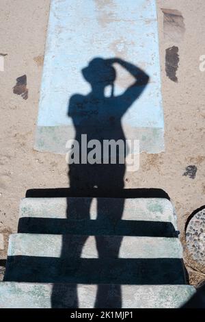 Un fotografo con un cappello fotografa la sua ombra cadendo su una scala, sabbia e cemento. Foto Stock