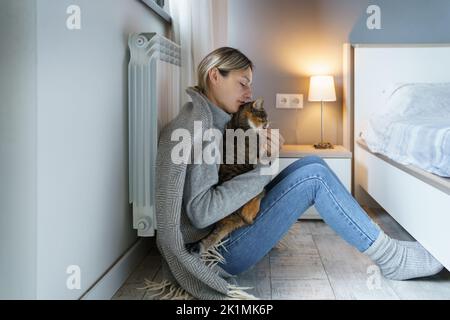 Una giovane donna coperta di coperta calda gioca con gatto che mangia animali con cibo seduto sul pavimento Foto Stock